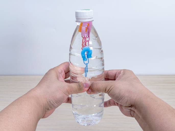 Two hands hold a clear plastic water bottle, partially filled with water, against a light wood-grain table and a plain white background. Inside the bottle, several small, looped pieces of colorful flexible straws are connected with paperclips, forming a chain-like structure that floats vertically in the water. The hands gently grip the bottle, likely as part of a science experiment or demonstration, possibly illustrating buoyancy or the Cartesian diver principle.