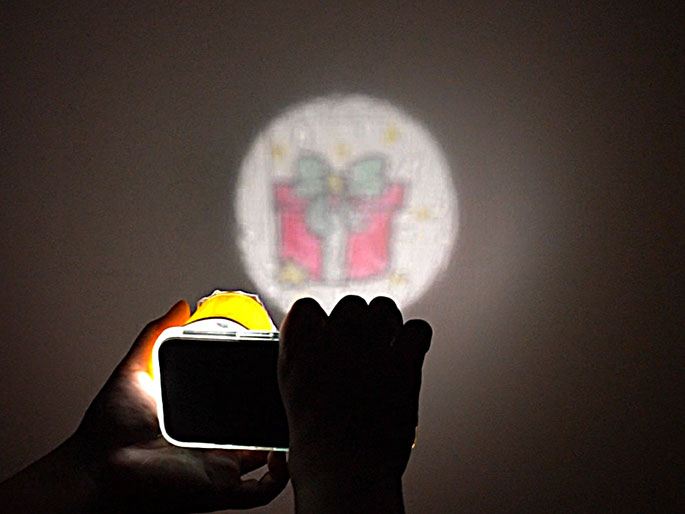 A hand holds a smartphone with a yellow attachment projecting a circular image of a Christmas present onto a dark surface, with another hand partially visible in the foreground.