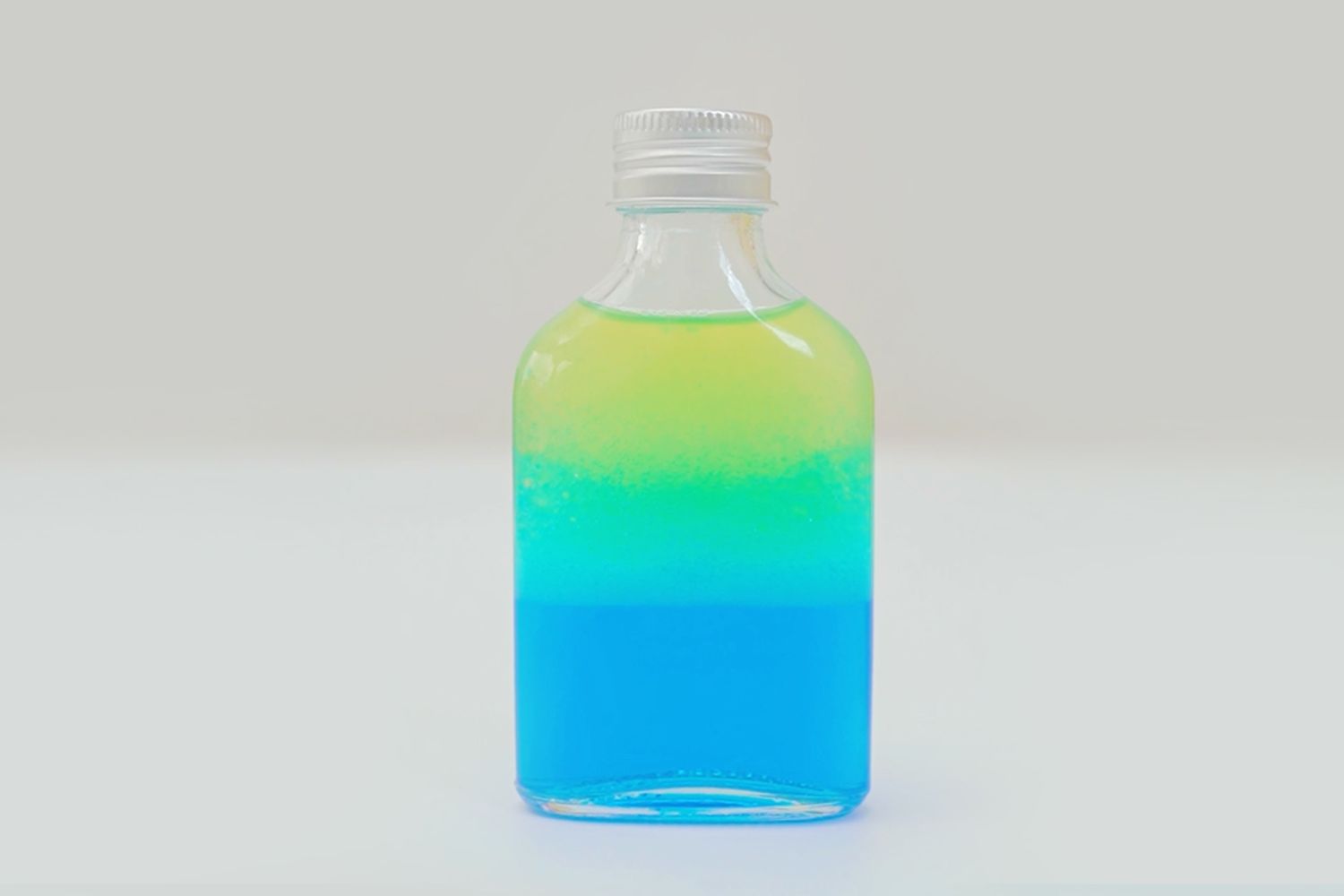 A small, rectangular glass bottle stands on a white surface against a plain, off-white background. The bottle is sealed with a silver screw-top lid and contains two distinct layers of brightly coloured liquid; a layer of pale yellow sits atop a layer of a more vibrant blue. There is a very slight mixing of the two colours where they meet, creating a thin, slightly green band. The bottle is positioned centrally, and the image is sharply focused, highlighting the clear separation and bright colours of the liquids. The overall impression is one of a completed sensory bottle, ready for use.