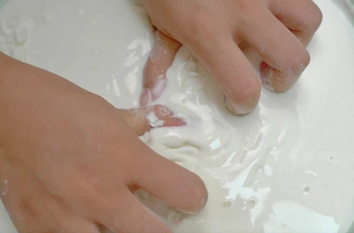 Crikey, look at that! A pair of hands are having a whale of a time playing with the cornflour slime, making it both solid and liquid all at once. It's a proper mind-boggling mess, innit? Such fun!