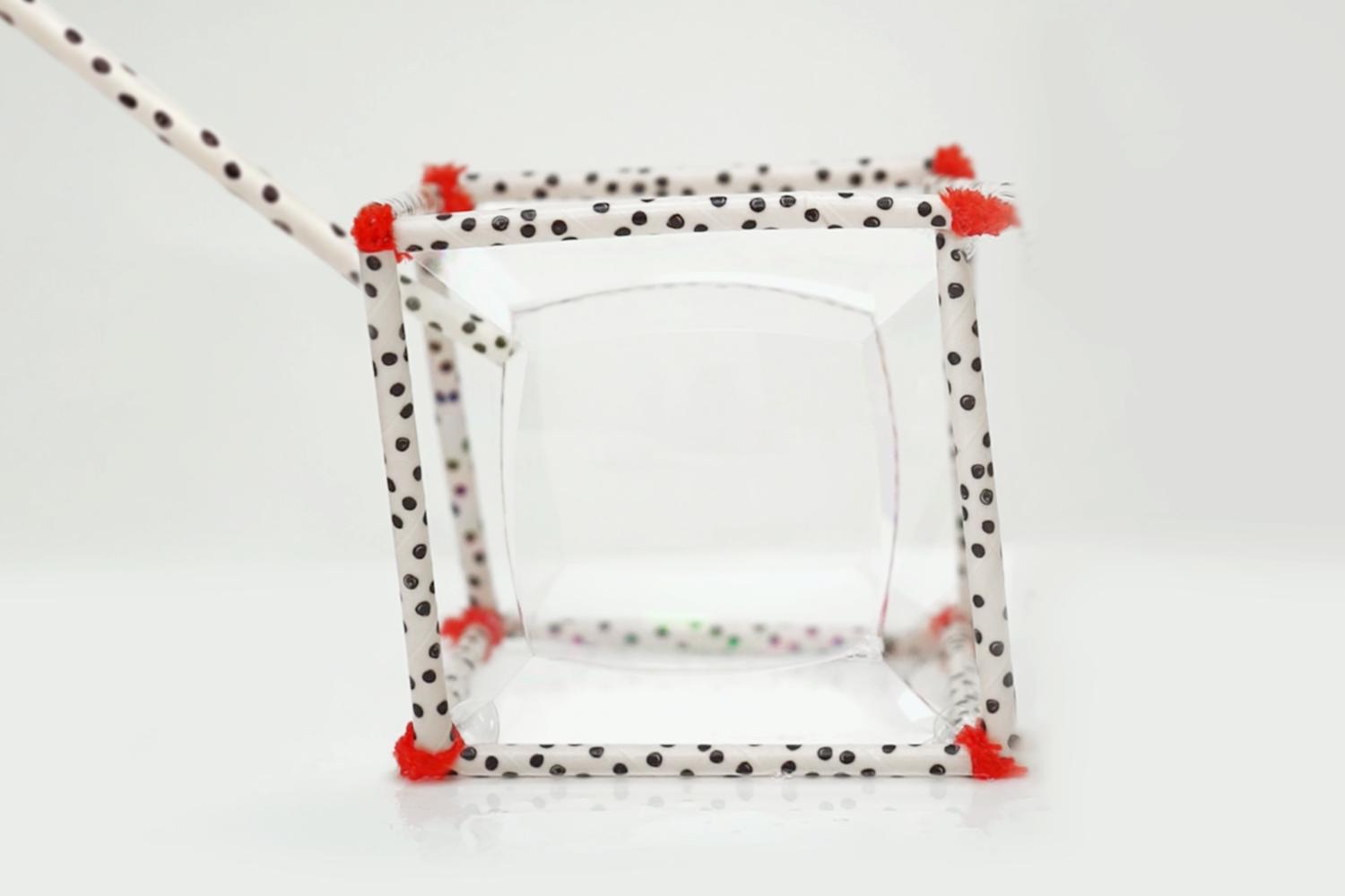 A cube-shaped frame made from black-polka-dotted straws, secured with red pipe cleaners, contains a clear soap bubble, with a straw extending from one corner.