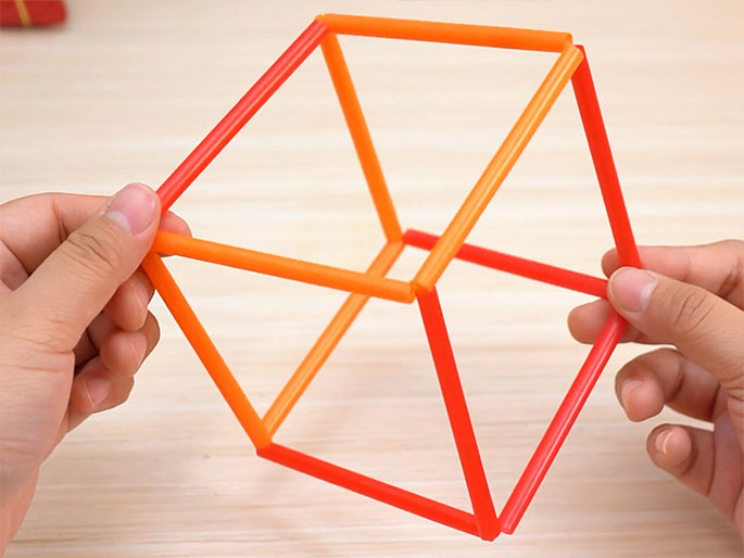 Two hands hold a three-dimensional cube constructed from short segments of orange and red drinking straws. The cube is held at a slight angle, displaying its form. The light wood-grain background provides contrast to the colorful straws. The image suggests a simple construction project or a geometric model made for educational or recreational purposes.