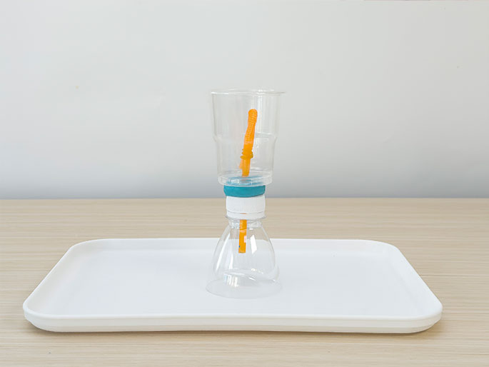A homemade contraption constructed from a clear plastic bottle and cup, connected by an orange straw, sits on a white tray against a plain white background. The bottle is inverted and attached to the cup, which is upright. A small amount of turquoise putty or clay is visible around the bottle's opening where it connects to the straw. The setup suggests a science experiment or DIY project, possibly related to air pressure or water flow.