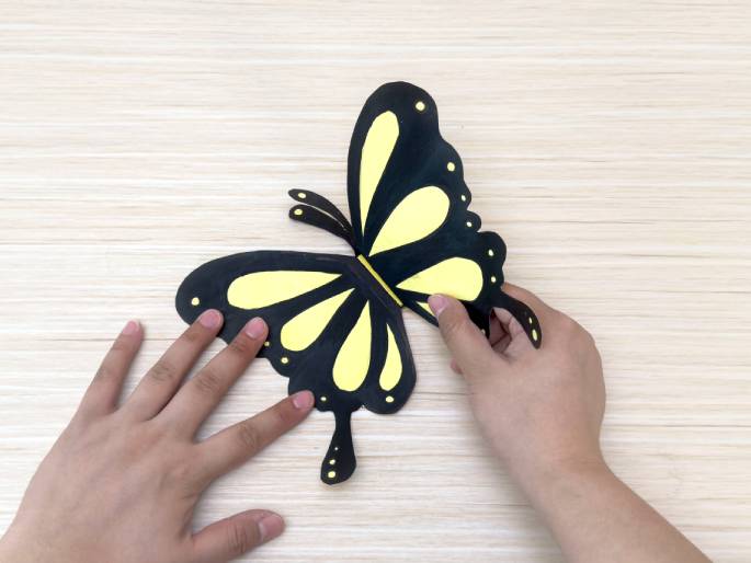 Two hands hold a completed paper butterfly craft against a light wood background. The butterfly, with black wings and yellow markings, is being held by its lower and upper wings. The wings are slightly raised, giving the butterfly a three-dimensional appearance. The image showcases the finished product of a simple paper craft project.