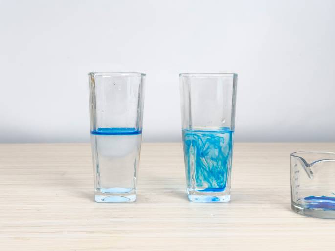 Two clear glasses partially filled with water and blue coloring sit on a light woodgrain table against a plain white background. In the left glass, the blue coloring has settled in a distinct layer at the bottom. In the right glass, the blue coloring is swirled and dispersed throughout the water, creating a marbled effect. A clear glass measuring cup with a wooden handle containing residual blue coloring sits to the right of the glasses. This image likely illustrates a density experiment or demonstrates the mixing of liquids.