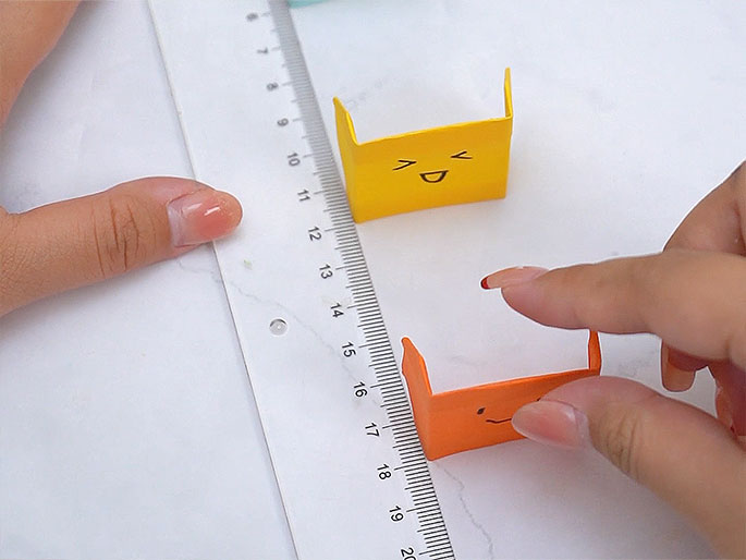 Two small, folded paper figures with drawn-on faces are positioned near a ruler on a white surface. The figure closer to the ruler is yellow with a happy expression, while the second figure, slightly further away and being held by fingers, is orange with a more neutral or slightly unhappy face. A person's hands are visible, one near the ruler and the other interacting with the orange figure. The ruler and the placement of the figures suggest they may be part of a measuring activity or a game involving distance and expressions.