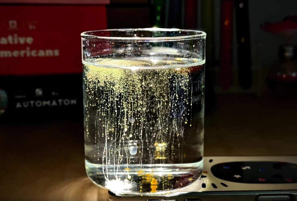 A clear glass filled with water sits on top of a smartphone. Tiny, gold-colored particles are slowly drifting down through the water, creating thin, shimmering trails. The smartphone's light illuminates the glass from below, enhancing the visual effect of the descending particles. The background includes books and test tubes, hinting at a learning or experimental setting.