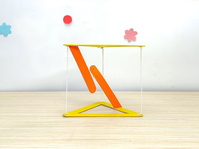 A three-dimensional craft object, constructed from yellow and orange craft sticks and white string, sits on a light wooden table. The base is a yellow triangle, and the string extends upwards to form a prism-like shape. Two orange sticks are suspended within the string structure, appearing to swing or hang freely. A white wall forms the backdrop, decorated with simple cutouts of a light blue flower, a red circle, and a pink flower.