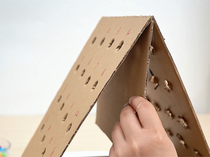 A small child interacts with a homemade game board constructed from cardboard. The board is triangular, resembling a slanted house, with numbered holes. Small, colorful balls and marbles are placed into the corresponding numbered holes. The child selects numbered cards and attempts to match them to the holes on the board. A small bowl of colorful gumballs sits beside the game. The video shows the child successfully placing some balls but also missing a few.