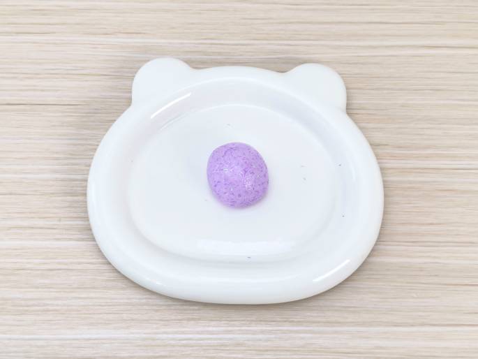 A small, oval-shaped piece of light purple slime or putty, speckled with glitter, rests on a white plate with bear-ear-shaped protrusions. The plate sits on a light woodgrain table. This image likely shows the finished product of a slime-making activity.