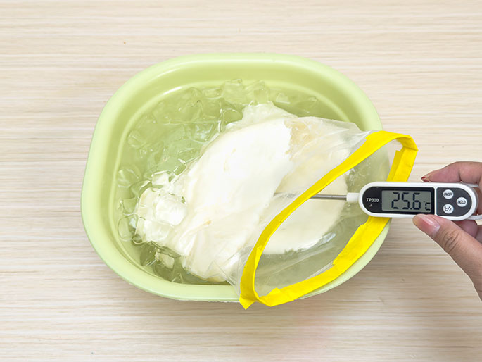 A sealed clear plastic bag with a yellow zip-top, containing a solid, off-white substance, is submerged in a light green bowl filled with ice water. A hand holds a digital thermometer; its probe is inserted into the bag, and the display reads 25.6 degrees Celsius. The image suggests a process of cooling or chilling a food item in an ice bath. The background is a light wood surface.