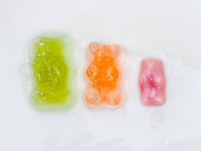 Three gummy candies sit on a white surface. From left to right, they are: a light green gummy bear that appears slightly melted, a translucent orange gummy bear, and a smaller, rectangular pink gummy candy that also appears somewhat melted. The image likely depicts a science experiment or demonstration involving the effects of heat or another factor on the candies.
