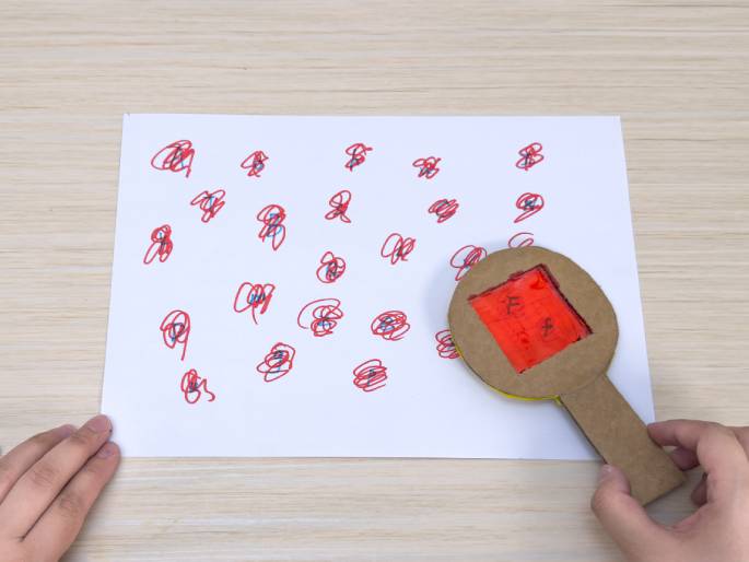 Two hands, a piece of paper, and a cardboard magnifying glass are displayed on a light wood-grain table. The paper is covered in red scribbles with small blue marks at their centers. The right hand holds the cardboard magnifying glass, which has a red-colored square window, over the paper. The left hand is positioned at the bottom left corner of the paper. The setup suggests a visual searching or decoding game using a homemade color filter.