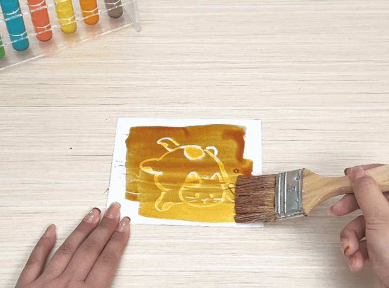 A hand is positioned to hold or adjust a blank white piece of paper on a light wood-grain table. In the top left corner, a rack of colorful test tubes is visible, suggesting a setting related to a science experiment, art activity, or educational demonstration.