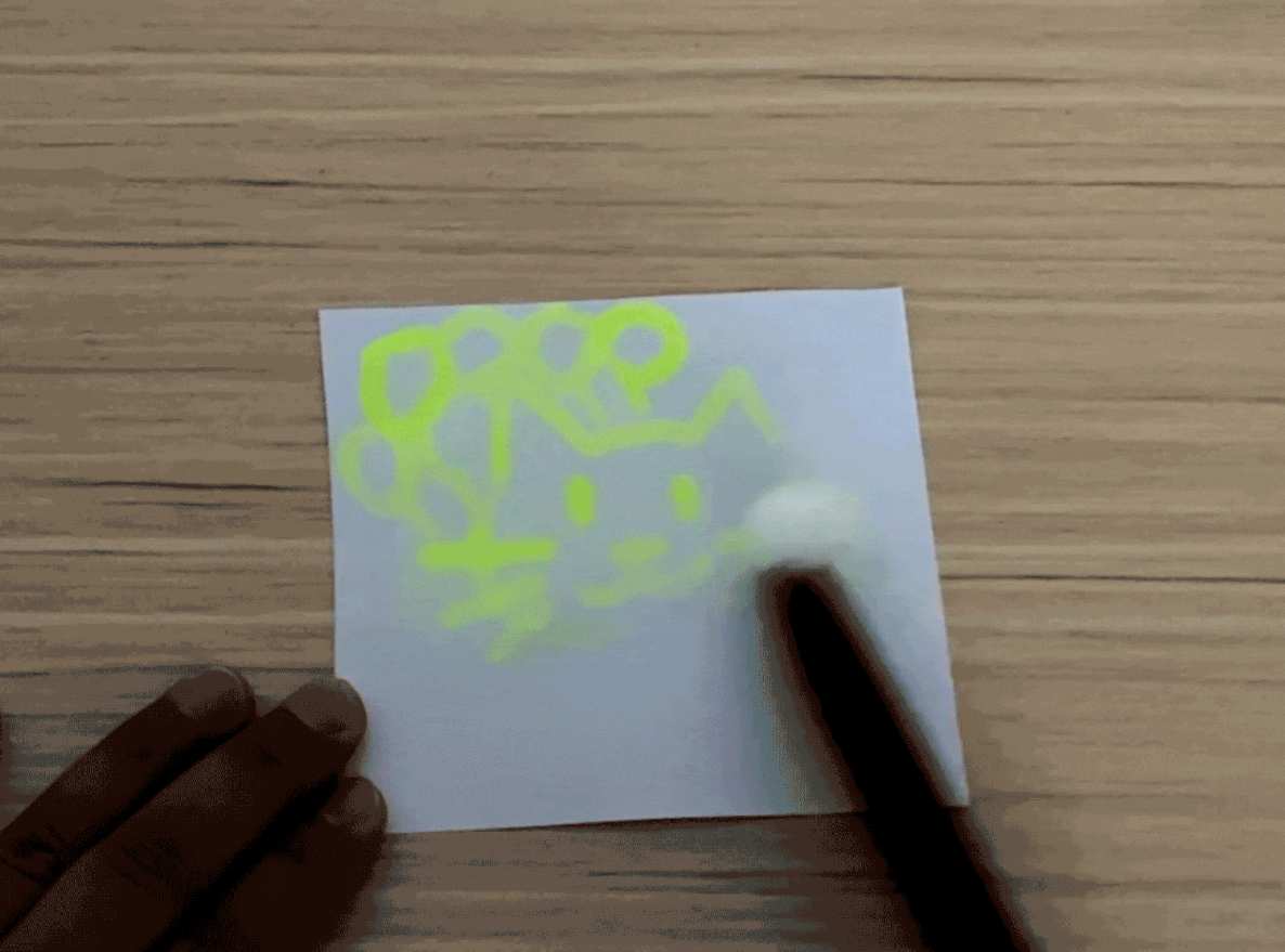 A hand partially visible in the bottom left corner holds a piece of white paper against a light woodgrain table. The paper has a few faint, light-colored markings on it, possibly from a liquid or light heat exposure. The focus is on the paper and its subtle markings, suggesting the result of an experiment or a creative process.