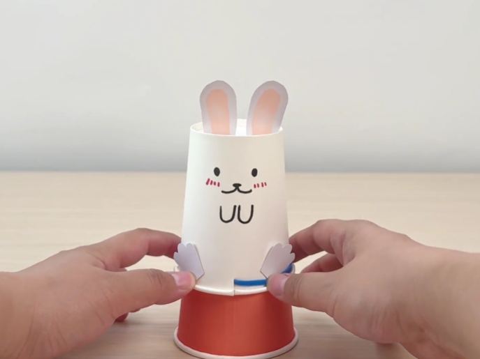 Two hands play with a small toy made from paper cups. The top cup is white and decorated with a cute, hand-drawn bunny face, complete with pink ears and small paws. A thin blue band is wrapped around the bottom of the white cup. The white cup sits on top of an orange paper cup. The hands gently push the sides of the white cup, causing it to pop up and off the orange cup, like a simple jumping toy. The video demonstrates a fun and easy craft activity using paper cups to create a small, jumping bunny toy.