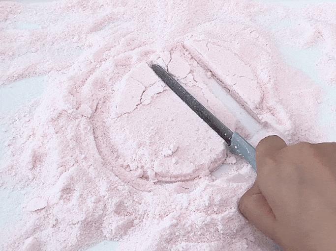 A hand uses a butter knife or small spatula to cut into a circle of light pink dough or mixture that is dusted with a similar light pink powder. The mixture and powder rest on a white surface. The scene suggests a baking or cooking activity.