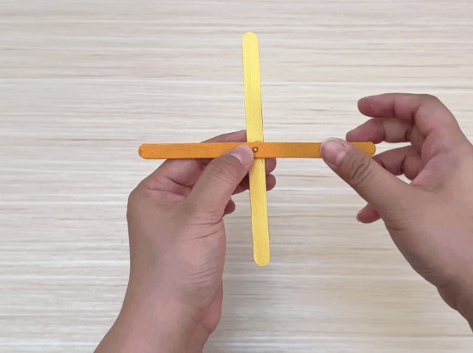 A hand holds a small, homemade toy resembling a whirligig or spinner. The toy appears to be constructed from a small, round object, possibly a bottle cap or button, attached to a light blue stick or handle. Yellow paper blades are affixed to the round object. A white string is tied to the handle and looped into a ring at the other end. The hand pulls the string back and forth, causing the yellow blades to spin rapidly, creating a blur of motion. The video demonstrates a simple DIY toy that can be made with readily available materials.