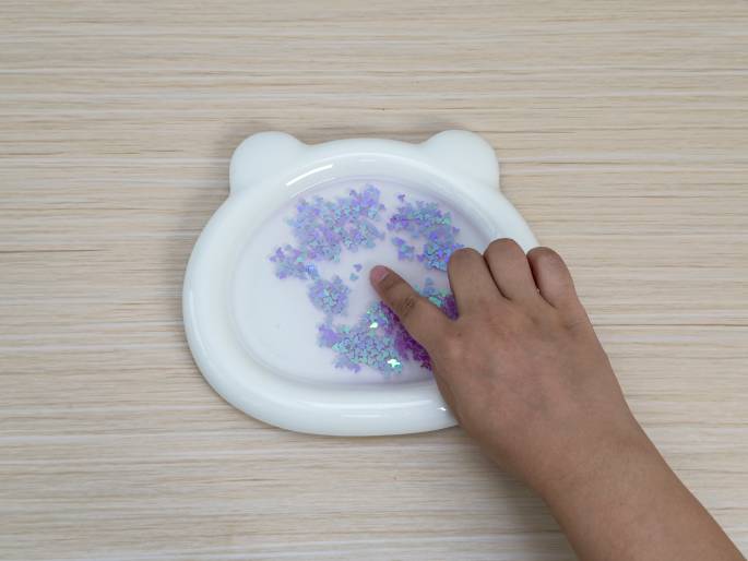 On a light wood-grain table, a white, bear-shaped dish holds water and light purple glitter. A hand interacts with the glitter, with a finger gently touching and seemingly moving the glitter within the water. The scene suggests a step in a craft activity, experiment, or sensory play.