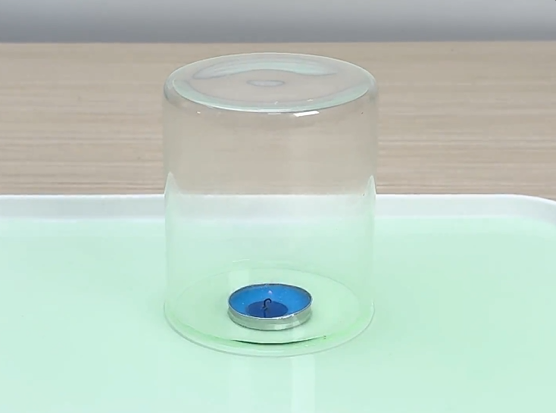 A small, lit tea light candle in a metal holder sits on a white tray filled with a light green liquid. A clear glass cylinder is placed over the candle. The candle flame burns steadily for a short time before gradually diminishing and extinguishing. The green liquid is then drawn up into the glass cylinder. This video demonstrates a classic science experiment illustrating the consumption of oxygen by fire and the resulting change in air pressure.