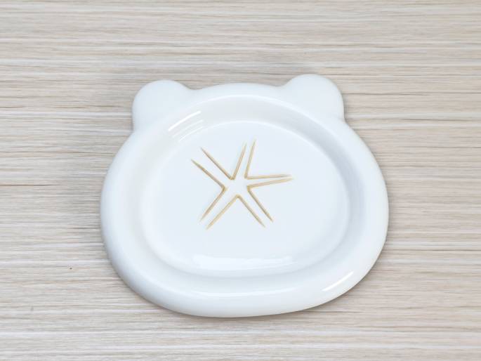 A white plate with small ear-like protrusions sits on a light wood-grain surface. Several toothpicks are arranged in a starburst or asterisk pattern in the center of the plate, suggesting the completion of a simple craft or arrangement.