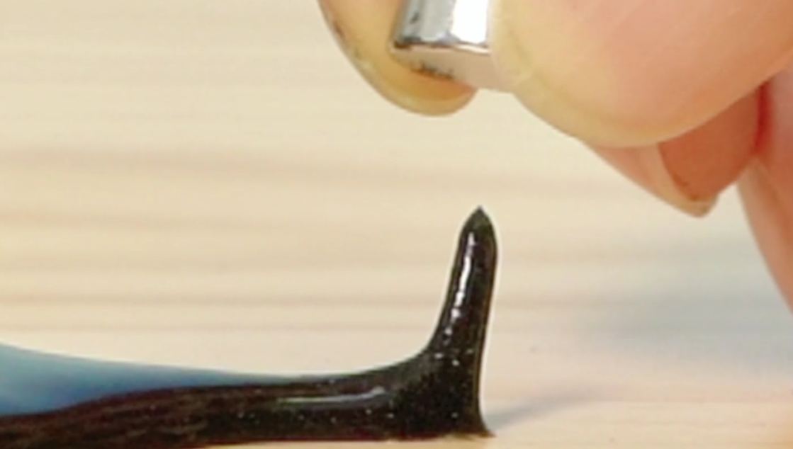 A dark-coloured slime, adorned with googly eyes, reacts to a magnet being held near it on a wooden surface.