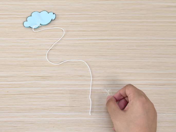 A light blue paper cloud cutout is attached to a white string that trails down onto a light wood-grain surface. A hand at the bottom of the frame holds a small piece of clear tape, preparing to attach something to the string. This image likely depicts a stage in a crafting or decorating project.