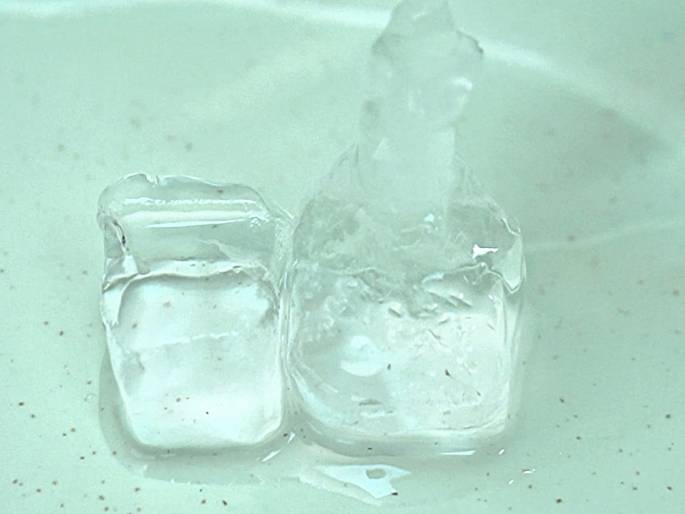Close-up of two melting ice cubes on a pale green surface. A small amount of meltwater has pooled around the bases of the cubes. The surface beneath them has small, dark speckles. One ice cube is more irregularly shaped, possibly due to faster melting or a previous break.