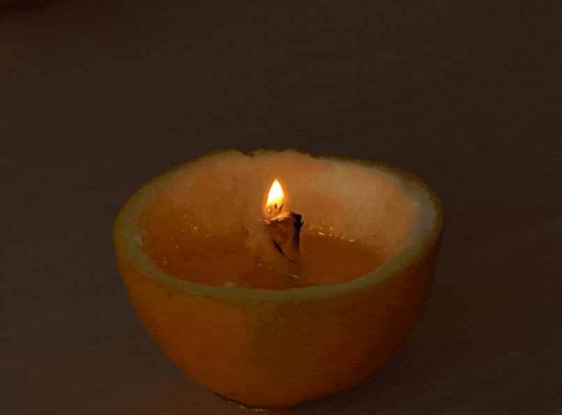 A small, makeshift candle is created using an orange peel as a container. The orange has been cut in half, and the pulp removed, leaving the rind. A small piece of wick or string sits in a pool of orange oil or juice inside the bottom half of the rind. A lit match or lighter ignites the wick, creating a small flame. The top half of the orange rind is then placed back on top, partially covering the flame and creating a small, enclosed orange candle. The video demonstrates a simple DIY project for creating a candle using an orange.