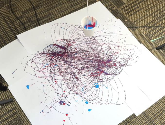 A small cup or container, likely punctured with holes, hangs suspended above a large piece of white paper. The cup contains red and blue paint, which drips and splatters onto the paper below. The cup swings in a pendulum motion, creating swirling and overlapping patterns of red and blue lines and splatters on the paper. The video captures the process of creating abstract art using a swinging pendulum and paint.