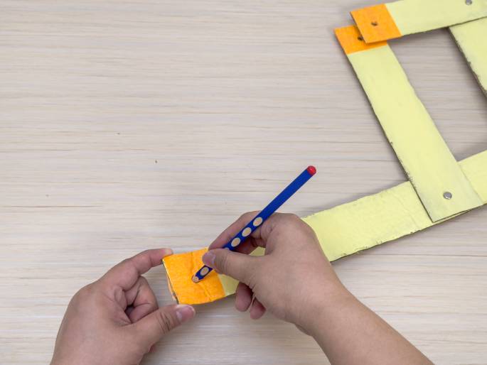 On a light wood grain surface, two hands are shown working with pieces of pale yellow cardboard, one of which has a bright orange section. One hand holds a smaller piece of cardboard with the orange section, while the other hand uses a blue pencil to make a mark on the orange area. In the background, more connected cardboard pieces, also partially orange, form an almost-square shape. This image appears to depict a step in a crafting project using cardboard.