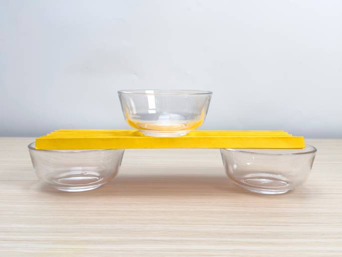 Three small, clear glass bowls are arranged on a light wood-grained surface. Two of the bowls are placed a short distance apart, and a third bowl is balanced on top of a folded piece of yellow paper that spans the gap between the lower bowls. The yellow paper acts as a bridge or platform. This arrangement likely demonstrates a physics or engineering concept related to weight distribution, balance, or structural strength.