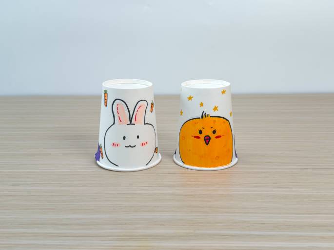 Two decorated paper cups sit on a light wood-grain table against a plain off-white background. The cup on the left features a drawing of a cute bunny rabbit with carrots, while the cup on the right has a drawing of a surprised-looking chick and stars. These cups appear to be part of a children's craft project.