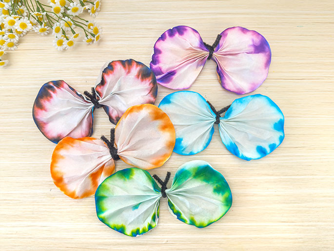 Several colorful coffee filter butterflies are arranged on a light wood surface. Each butterfly is made from two round filters, folded and tied together in the center with a black pipe cleaner to create wings. The filters are decorated with vibrant tie-dye patterns in various colors, including orange, pink and dark purple, blue, and green. A small bunch of daisies is positioned in the top left corner of the image.