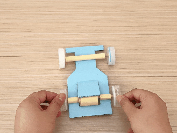 A small, light blue toy car, crafted from cardboard and plastic bottle caps for wheels, rests on a light wooden table. The car features exposed axles made from what appears to be rolled-up paper or thin cardboard tubes. A hand briefly enters the frame at the bottom right. The car does not move, and a popping sound is heard, possibly indicating an off-screen action or the sound of something being placed on the table.