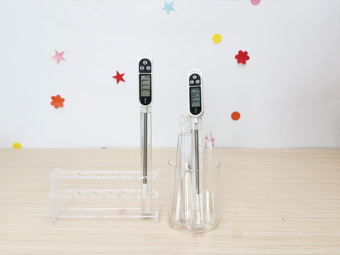 Two digital thermometers are shown inserted into glass tubes containing clear liquid. One thermometer sits in a test tube held by a clear acrylic rack on a light wood-grain table. The other thermometer is placed in a glass beaker containing several other glass tubes, also filled with liquid. The thermometers display different temperature readings. The background is a white wall decorated with colorful star and flower stickers.
