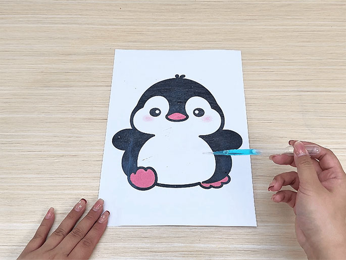 A hand holds a pipette filled with blue liquid over a drawing of a cartoon penguin on white paper. The penguin's outline is black, its feet are pink, and some gray crayon shading is visible on its body. Another hand is partially visible at the bottom left, holding the paper. The background is a light wood surface. The image appears to depict a child's art project in progress, using a crayon resist technique with liquid watercolor.