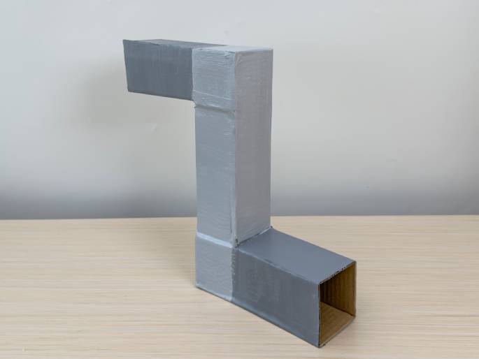 A finished, gray-painted cardboard structure resembling a periscope or pipe sits on a light wood-grain table. The structure is L-shaped, formed by two rectangular boxes joined at a right angle. One end of the horizontal section is open, revealing a square opening. The object stands against a plain white wall.