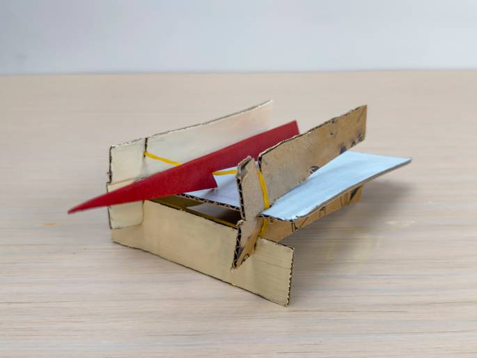 A small airplane or glider, crafted from various pieces of cardboard, sits on a light wood-grain surface. The fuselage appears to be made of plain, light-colored cardboard, while the wings are constructed from different types of cardboard: one is light grey or blue, and the other has a printed design. A bright red piece of cardboard, likely representing the tail fin, extends prominently from the rear. Yellow rubber bands are visible, likely used to hold parts of the plane together. This image showcases a completed DIY project, possibly a toy airplane.