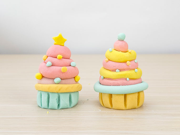 Two small decorative items made of modeling clay sit on a light woodgrain surface. The first resembles a Christmas tree-shaped cupcake. It has a light green base, a yellow cupcake wrapper, pink swirled frosting decorated with small colorful dots, and a yellow star on top. The second resembles a traditional swirled cupcake. It has a yellow base, a light green cupcake wrapper, and pink and yellow swirled frosting topped with a light green ball and small colorful dots.