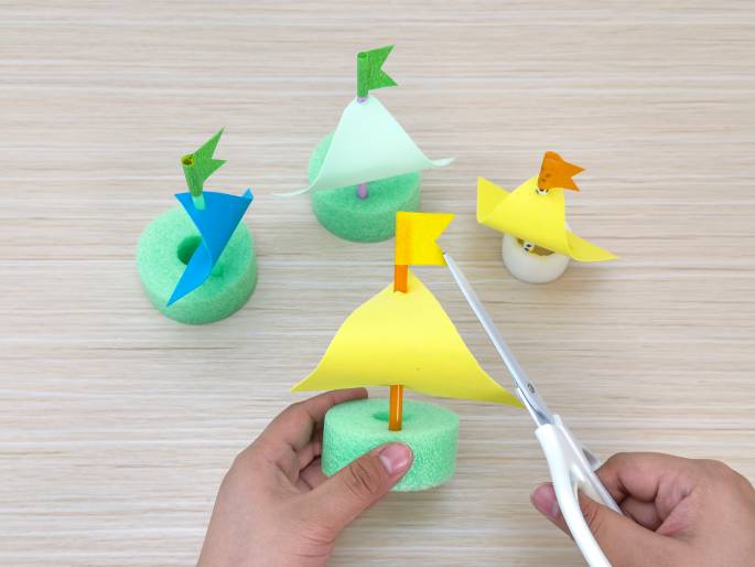 Several small, handmade sailboats are displayed on a light wood-grain table. Each boat has a light green foam ring for a hull and a colored paper sail attached to a straw mast with a small paper flag at the top. The sailboats have sails and flags in various colors: light green with a dark green flag, bright yellow with an orange flag, and blue with a light green flag. Two hands are shown in the foreground, one holding a yellow-sailed boat and the other holding a pair of white scissors, seemingly about to trim the sail. This image likely depicts a craft activity involving making toy sailboats.