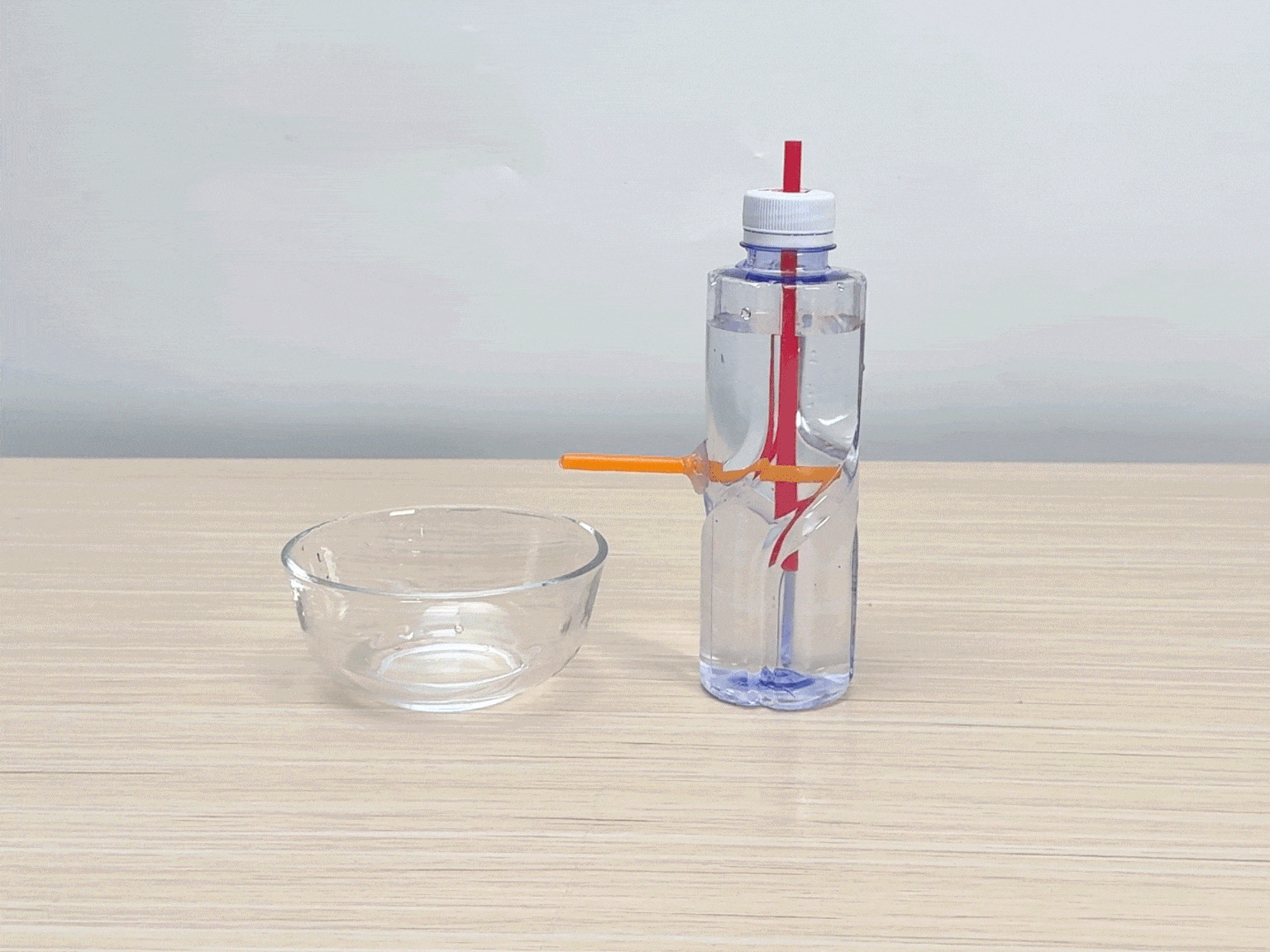 A clear plastic bottle, partially filled with water, stands on a light wooden table next to an empty clear glass bowl. A red drinking straw is inserted into the bottle's opening. An orange straw is inserted horizontally through the side of the bottle. A hand gently squeezes the bottle. Water flows out of the orange straw and into the bowl. The video demonstrates a simple science experiment, likely illustrating the principles of air pressure and siphoning.