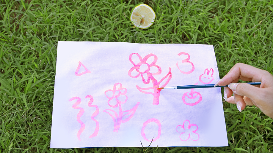 Ooh, look at that, a bit of artistic magic! A paintbrush is adding some lovely pink shapes to the paper – flowers, swirls, and numbers. It’s all rather pretty, innit? A proper bit of creative fun, made even better by the lovely green grass in the background. What a jolly scene!