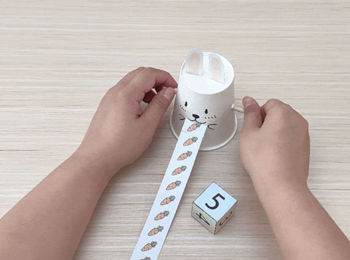 A paper cup, decorated with a cute bunny face and ears, sits on a light wooden surface. A long strip of paper, printed with small carrot images, is being pulled out from the bunny’s mouth. Small wooden sticks protrude from the sides of the cup, likely serving as arms. This video likely demonstrates a simple craft or toy, possibly designed for children, showcasing a bunny “eating” a paper strip of carrots.