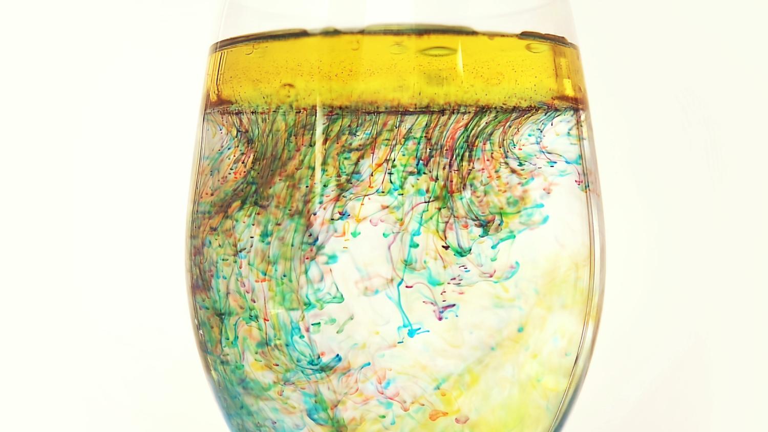 A wine glass is partially filled with water, overlaid with a layer of yellow liquid, and streaks of colourful inks create a marbled, rainbow effect.