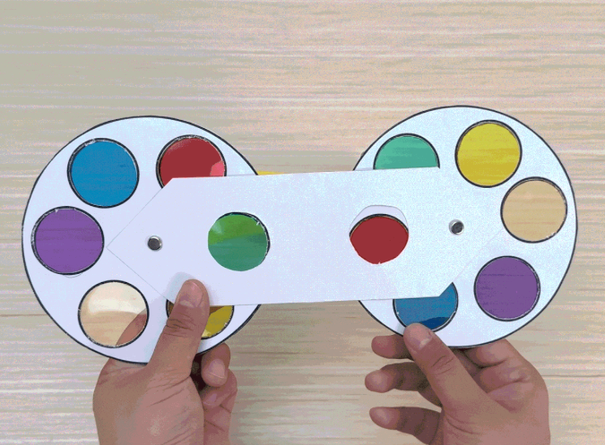 Two hands hold a completed paper color wheel device against a light wooden background. The device consists of two circular color wheels connected by a white paper handle. Each wheel has segments of translucent colored cellophane in red, yellow, green, blue, purple, and peach/orange. The handle has two red circles visible, suggesting a mechanism for viewing color combinations. A small brad or fastener is visible at the center point where the handle joins each wheel. This image showcases the finished color-mixing tool.