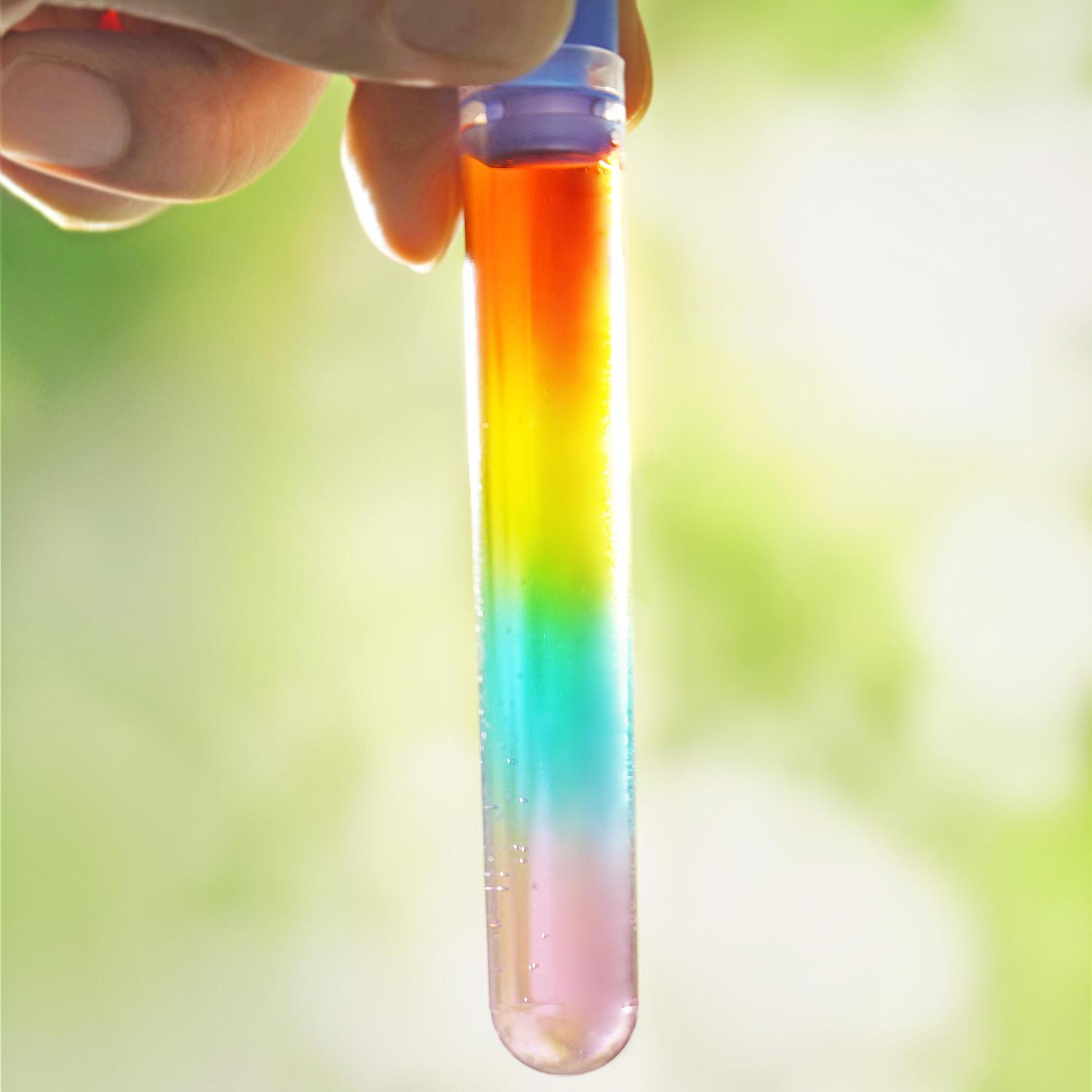 A hand gently holds a test tube filled with a layered liquid, creating a vibrant rainbow effect. The layers are clearly defined, showing distinct colours from top to bottom: red, orange, yellow, green, and pink. The test tube is transparent, allowing the colours to be seen clearly. The background is a blurred, out-of-focus image of green and yellow tones, providing a natural and pleasing contrast to the bright colours of the liquid. The image suggests a successful science experiment demonstrating liquid density and colour mixing.
