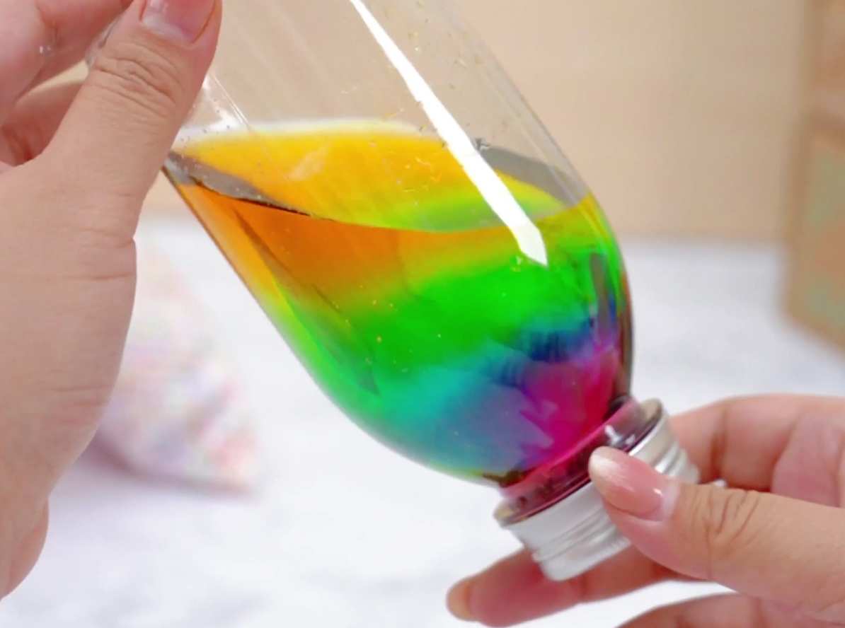 A pair of hands holds a clear plastic bottle filled with liquids forming distinct horizontal layers in rainbow colors. The layers, from top to bottom, are orange, yellow, green, blue, indigo, and violet. The hands gently tilt and rotate the bottle, causing the colorful liquids to swirl and mix slightly, demonstrating the different densities and viscosities of the liquids.