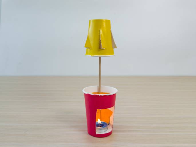 A small, homemade hot air balloon spins above a lit tea candle. The balloon is made from a yellow paper cup with cutouts, attached to a stick inserted into a red paper cup. The tea candle sits inside the red cup, providing the heat source. This demonstrates a simple science experiment.