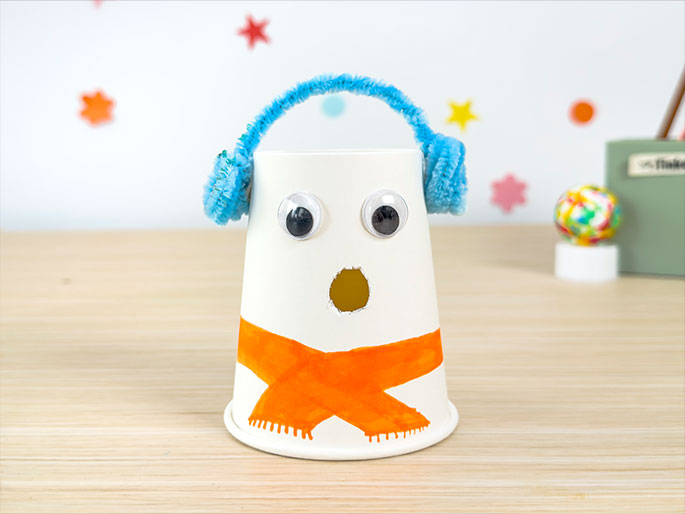 A pair of hands presents a snowman craft made from a white paper cup. The cup is decorated with googly eyes, an orange marker-drawn scarf, and blue pipe cleaner earmuffs. A small hole cut into the cup serves as the snowman's mouth. The background features a blurred assortment of colorful craft supplies and other finished projects. Bubbles float gently in the foreground, adding a whimsical touch.